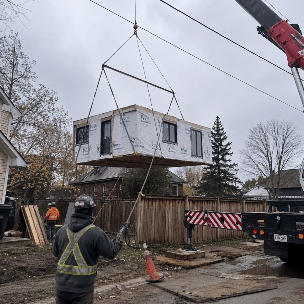 Prefab cottages Orangeville module being craned into a residential backyard during installation, with a mobile crane, lifting straps, utility lines and installation crew preparing to set the factory-built home