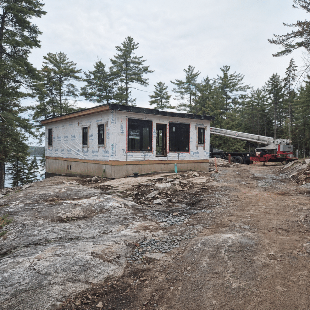 Cost to build a cottage in Muskoka: prefab cottage module on a foundation over exposed Canadian Shield rock with a crane still on site.